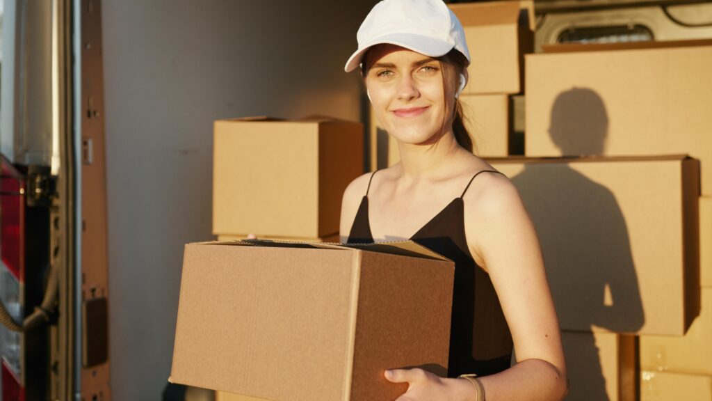 Photo of a girl holding moving boxes, standing with a smile as she carries a stack of packed boxes. The image highlights her readiness and enthusiasm for the moving process, emphasizing the personal and organized approach of the moving company in handling belongings.